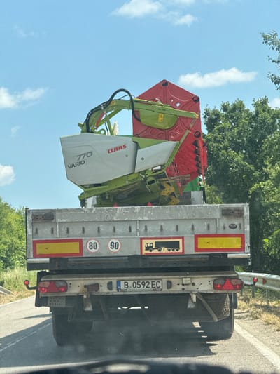 The image shows a close-up view of a trailer truck carrying agricultural machinery. The machinery, primarily green and red, appears to be a CLAAS 770 VARIO combine harvester. The truck is parked on the side of a road, with a highway barrier visible in the background, along with trees and a blue sky with some clouds. The time of day seems to be daytime, likely late morning or afternoon, given the bright sunlight casting shadows. The weather appears to be clear and sunny.
Visible text on the machinery includes "CLAAS" and "770 VARIO". On the back of the trailer, there are several safety markers, including speed limit signs (80 and 90) and reflective tape. A license plate is also visible, reading "B.0592 EC" with "BG" indicating Bulgaria. The truck itself is a flatbed trailer. The overall scene suggests the transport of agricultural equipment, possibly to or from a farm or for maintenance. The image is taken from a slightly angled perspective, with the truck positioned towards the right side of the frame. The condition of the machinery appears used, with some dirt and dust visible, which is common for agricultural equipment.