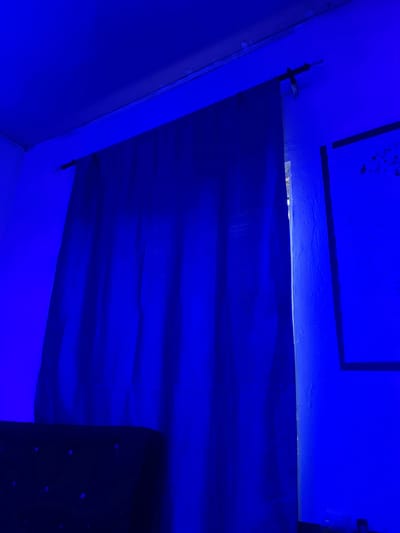 This media file depicts an interior scene bathed in intense blue light. The primary subject is a wall covered with a dark blue fabric, which appears to be draped or hung. The fabric has visible wrinkles and creases, creating horizontal lines across its surface. Above this fabric, another section of the wall is visible, painted a lighter shade of blue with some texture and what appear to be dark, rectangular elements or markings. To the left of the frame, a corner of the room is visible, revealing a white wall meeting a blue wall at an angle. A black clip or bracket is attached to the junction of the blue wall and the fabric, suggesting the fabric is secured in place. In the lower right corner, a portion of a dark object with scattered light-colored spots is visible, possibly a piece of furniture or another fabric.

The scene is artificial due to the pervasive blue lighting, making it difficult to determine the time of day or weather conditions. The environment appears to be indoors, possibly a room, studio, or a temporary setup. There are no people visible in the frame. The overall impression is one of a staged or decorated space, with the blue light creating a specific mood, potentially dramatic or atmospheric. No text is visible in the image. The location context provided, Pantinapu, Nigeria, does not offer any visual cues within the image itself.