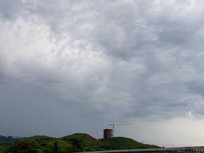 A drone shot captures a dramatic cloudy sky over Buknari, Georgia. The sky is filled with an expanse of gray, turbulent clouds, with some lighter patches suggesting breaks in the overcast. In the mid-ground, rolling green hills dominate the landscape. On the crest of one hill stands an unfinished concrete building, its skeletal frame exposed. A tall yellow construction crane is positioned next to the building, its arm extended horizontally. The foreground shows the dark silhouette of a railing, suggesting the vantage point is from a raised structure, possibly a bridge or elevated walkway. The overall scene evokes a sense of ongoing development against a backdrop of imposing weather.