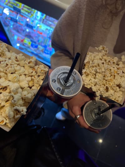 The image shows two individuals, seemingly at a movie theater in Rishon LeZion, Israel. Each person has a container of popcorn and a drink with a straw. The background includes what appears to be a vending machine with brightly colored items. The scene is dimly lit.