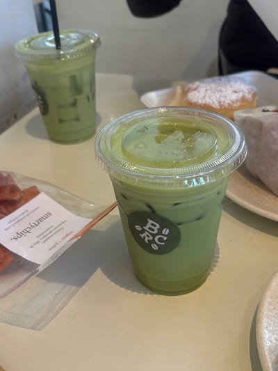 The image captures a close-up, overhead view of two iced matcha lattes on a light-colored table, likely at a cafe in San Pedro Garza García, Mexico. The latte in the foreground is in a clear plastic cup with a domed lid, featuring ice cubes and a vibrant green liquid. A circular green sticker with white lettering and symbols, possibly a cafe logo, is adhered to the cup. A plastic straw is inserted into the lid. Behind it, another similar iced matcha latte is visible, also in a clear plastic cup with a straw. To the left of the foreground latte, a clear plastic bag contains what appear to be dehydrated fruit chips, with a white label featuring black text. Further in the background, a pastry dusted with powdered sugar is partially visible on a white plate, and a blurred figure in dark clothing is present. The overall impression is of a casual cafe setting with refreshing beverages and snacks.