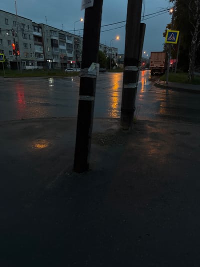 This media file depicts a wet street scene at dusk in Nevyansk, Russia. The perspective is from ground level, looking towards a city street with a multi-story apartment building on the left and a road on the right. The road surface is wet and reflective, indicating recent rain, with streaks of light reflecting from streetlights and vehicle lights.

In the foreground, two dark wooden utility poles are prominently visible, extending vertically from bottom to top of the frame. The one on the left is partially obscured by the edge of the image, while the one on the right is more centered.

Further down the street, there's a pedestrian crossing sign with a yellow background and a blue symbol of a person walking. A truck with "SANDVIK LIVE" written on its side is visible on the road, its brake lights and taillights illuminated. Traffic lights are also visible, showing a red signal. The apartment building on the left has numerous windows and balconies, with some lights on inside. Several streetlights cast an orange glow onto the wet road.

The overall mood is somewhat somber and quiet, likely due to the weather and time of day. The reflections on the wet pavement add a sense of depth and atmosphere to the scene. The time of day is clearly dusk or early evening, as evidenced by the low light and the illumination from artificial sources.