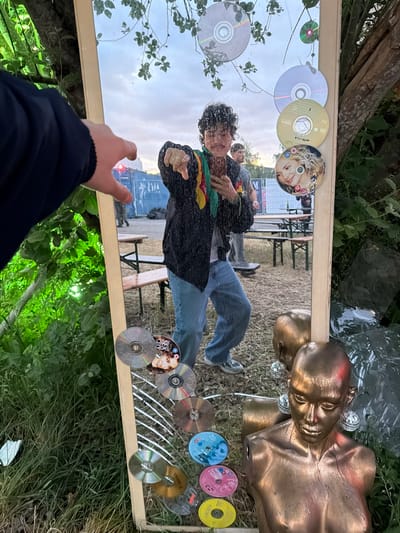 The scene is set outdoors in Copenhagen, Denmark, featuring a large, vertically oriented mirror adorned with numerous compact discs (CDs) and DVDs. The mirror has a light-colored wooden frame and reflects an outdoor gathering.

In the reflection, a young adult with dark, curly hair, wearing a dark jacket over a light shirt, blue jeans, and sneakers, is centered. They hold a smartphone in their left hand, seemingly taking a photo of themselves in the mirror, and extend their right arm, pointing an index finger outwards towards the viewer with a slight smirk. A colorful, striped fabric is draped over their left shoulder. The background of the reflection shows a light-colored sky with clouds, green foliage, several wooden picnic tables and benches on dry, grassy ground, and a long blue temporary fence or wall in the distance. Other individuals are visible walking among the tables in the reflected background. A red light source is discernible in the upper left of the reflection. Among the discs attached to the mirror are a gold-colored Metallica CD/DVD, and a "Sex and the City" DVD featuring Sarah Jessica Parker. Other discs are generic silver, blue, pink, and yellow.

In the foreground, outside the mirror, the left arm and hand of the person taking the photograph are visible, clad in a dark blue long-sleeved garment, reaching towards the mirror's left edge. To the lower right of the mirror, two bronze-colored female mannequin busts are positioned. One is a partial, headless torso, while the other is a full head and upper torso, adorned with small, spherical disco ball earrings. The surface of this bust appears damp or shiny. The ground around the mirror consists of dry grass and dirt, with green foliage and tree trunks visible behind and to the left of the mirror frame. Some white debris is visible on the ground to the far left.