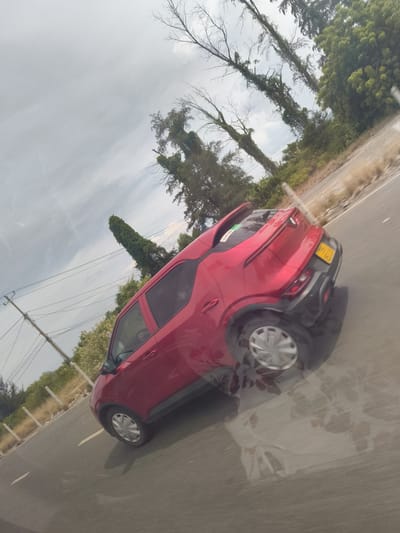 A red Nissan Juke, license plate 43A-161.70, is traveling on a paved road. The car is angled with its rear to the viewer, and its wheels are spinning, indicating motion. The road has yellow lane markings and is lined with low-lying vegetation and fences. In the background, there are trees and utility poles with numerous wires. The sky is overcast with some wispy clouds. The image has a slight tilt to the right, making it appear that the scene is at an angle.