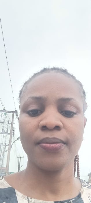 An adult female, characterized by a medium-dark complexion and braided hair, is depicted in a close-up, head-and-shoulders view. Her dark eyes are directed towards the viewer, and her full lips maintain a neutral expression. She is wearing a patterned garment featuring light and dark tones. The outdoor background consists of an overcast, light grey sky. To the left, a utility pole structure with multiple power lines and a transformer is clearly visible, characteristic of urban infrastructure. Additional utility poles and faint distant structures are present further back. The scene is located in Aba, Abia, Nigeria.