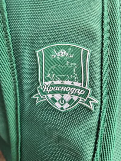 This is a close-up, eye-level shot of a green embroidered crest on a green fabric material. The crest is oval-shaped and outlined in white. Inside the crest, there are various green and white emblems. On the left side, there appears to be a silhouette of a bull or a cow, and to the left of that, a circular emblem with what looks like crossed flags or sticks. A banner curves over the top of the crest with white lettering on a green background. The letters are stylized and appear to read "Kpacнодар" which is Cyrillic for Krasnodar. The right side of the crest has a checkered pattern with a white outline of what could be a horseshoe shape. The fabric material has a visible woven texture. The lighting suggests it is daytime. There are no people or other objects visible in the frame. The location context is Lenina, Russia.