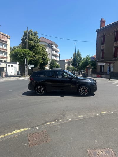 A black BMW X5 SUV is driving through an intersection in Clermont-Ferrand, France. The car is in the middle of the road, turning left. The sun is shining, and the sky is clear. There are buildings on either side of the road, as well as trees and street signs. One street sign points to "CHAMALIÈRES" and another to "CLERMONT-FP-CENTRE SALINS". The road is paved and has white lane markings. There is a man driving the car.
