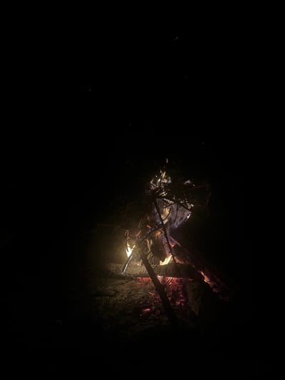 The image depicts a campfire in a dark environment. The fire is contained within what appears to be a natural or man-made fire pit, surrounded by earth and logs. Flames and glowing embers are visible, illuminating the immediate area around the fire. Thin branches and other smaller pieces of wood are burning. The scene is predominantly dark, with the fire acting as the main light source. No people are visible. The location is Юзуфоўскі сельскі Савет, Belarus.