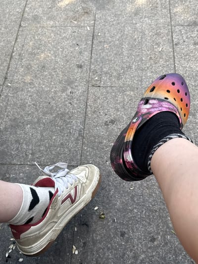 The image shows two feet resting on a gray, paved surface, likely a sidewalk or plaza, with scattered white and green debris. The left foot is wearing a white Vans sneaker with red accents and a white sock with black chevron patterns. The right foot is wearing a colorful Croc with a sunset gradient pattern, black socks with a white and black striped cuff. The scene appears to be outdoors. The location specified, Il’inskiy, Russia, is not visually discernible from the image itself.