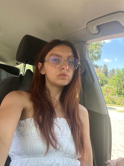 A young woman with long, auburn hair and glasses is pictured sitting in the passenger seat of a car. She is wearing a white, strapless top. The interior of the car is visible, including the headrest and a grab handle. Through the open car window, a rural landscape with green trees and a blue sky with some clouds is seen. The background suggests a sunny day, likely in the city of Tryavna, Bulgaria, given its characteristic mountainous terrain and greenery. There are no other people or significant objects visible in the scene.
