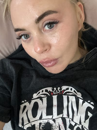 A close-up, angled shot features a fair-skinned individual with light blonde hair, light eyes, and full, glossy lips. Their makeup includes dark eyebrows and enhanced eyelashes. They are wearing a dark grey hooded sweatshirt. The sweatshirt has a white graphic on the front. The background appears to be a light pink or beige surface, possibly bedding or a pillow. There is no information available in the image about the city of Высокий, Russia.