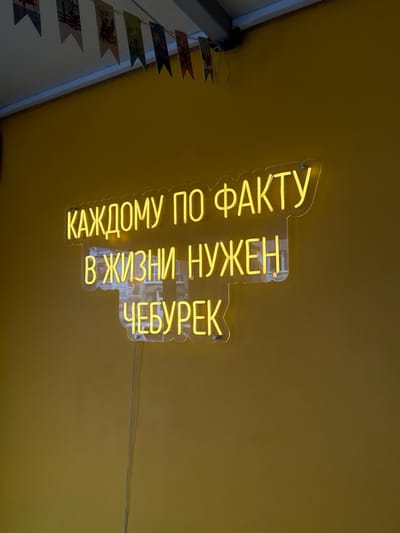 The image shows a yellow neon sign on a yellow wall. The sign reads "КАЖДОМУ ПО ФАКТУ В ЖИЗНИ НУЖЕН ЧЕБУРЕК", which translates to "EVERYONE NEEDS A CHEBUREK AS A MATTER OF FACT IN LIFE". The sign is mounted on a clear acrylic backing and illuminated with yellow neon light. There is a string of small decorative flags hanging from the ceiling above the sign. The wall appears to be an interior wall, possibly in a cafe or restaurant. The image does not provide any specific geographical information, so the location in Ussurijsk, Russia cannot be determined. There are no people or actions depicted in the image.