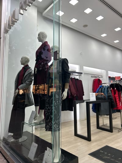 This image captures an interior view of a contemporary clothing store, likely a display window or a section of the sales floor, located in Koridallos, Greece. The scene features three full-body mannequins positioned behind a reflective glass barrier, showcasing winter or autumn attire.
From left to right (as if the image were rotated upright), the first mannequin wears a grey top with red accents and dark trousers. The middle mannequin is dressed in a dark maroon patterned top with dark trousers. The third mannequin, positioned slightly higher and further back, sports a dark coat with a visible beige sleeve over dark clothing. A small electronic device, possibly a smartphone, rests on the glass display near this mannequin.
In the background, clothing racks are visible. One rack holds a selection of puffer jackets and vests in red, blue, and black. Another rack displays dark red or maroon garments. A small white sign on a rack indicates a sale with percentages such as "-30%, -40%, -50%." The store's interior features light-colored wooden flooring, white walls, and a white ceiling illuminated by numerous square recessed lights. Significant reflections on the glass barrier create visual distortions of the mannequins and the surrounding store environment. A dark floor mat with white text, reading "MAKI Y..." and a signature-like design, is partially visible on the right side of the image. The setting suggests a well-lit retail space dedicated to fashion display.