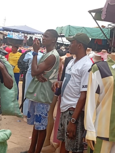 An outdoor market in Asaba, Nigeria, is observed under an overcast sky. Two adult males stand prominently in the foreground amidst numerous clothing mannequins. The male on the left, wearing a light green sleeveless top, distressed blue denim shorts, and a blue wristwatch, holds a red object to his chin. The male on the right wears a dark green baseball cap, a white t-shirt with a partial blue design and "ARF" text on the sleeve, distressed grey denim shorts, a dark-faced wristwatch, and a ring; a necklace is also visible. Both men look toward the right of the frame. Mannequins, predominantly headless torsos of varying skin tones, display diverse garments including shirts and dresses. In the background, a woman in a yellow shirt carries multiple bottled waters in a black basin on her head. Market stalls with blue and green tarpaulin coverings are visible, as are large green sacks on the left. The ground is composed of unpaved earth.