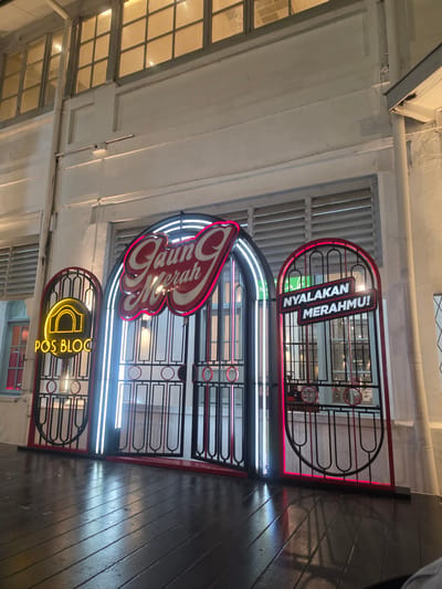 The image shows a neon sign above an arched entrance in the city of Medan, Indonesia. The sign reads "Gaun Merah" in cursive script with red neon outlines and white fill. To the left of the main entrance is a smaller arched sign with a yellow neon outline and a stylized depiction of a building, with the text "POS BLOC" below it. To the right of the main entrance, there is another arched gate with a sign that reads "NYALAKAN MERAHMU!" in white lettering against a red neon outline. The signs are illuminated with neon lights, casting a glow on the surrounding white wall and the dark wooden floor. The overall scene suggests a commercial establishment, possibly a restaurant or event space, with a vibrant and retro aesthetic. The architectural style of the building, with its large windows and decorative elements, hints at a historical building.