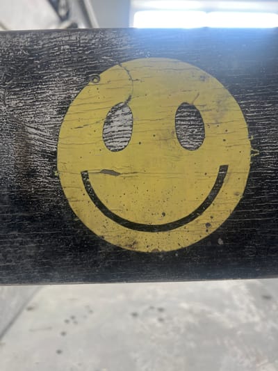 The image is a close-up shot of a weathered yellow smiley face painted on a dark, textured surface, possibly wood or metal. The smiley face is imperfect, with areas of paint chipped and faded, revealing the darker material underneath. The eyes are simple black ovals, and the mouth is a curved black line. The background is out of focus, showing hints of a bright, possibly overcast sky or artificial lighting. The lower portion of the image shows a blurry, light-colored surface with a scattering of dark spots. No specific location in Тэльмінскі сельскі Савет, Belarus, can be identified from the visual information provided. There are no people or discernible actions taking place.