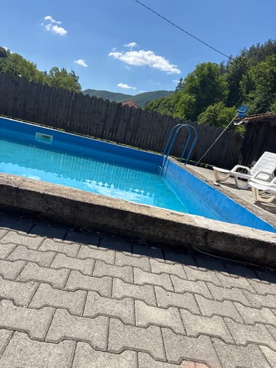 The image shows an outdoor swimming pool located in Liava Reka, Bulgaria. The pool is rectangular with bright blue lining and filled with clear blue water. A metal ladder with blue steps is attached to the side of the pool. The pool area is paved with gray interlocking concrete pavers. To the right of the pool, two white plastic lounge chairs are visible. Behind the pool, there is a wooden fence and a dense green forest covering rolling hills under a bright blue sky with scattered white clouds. A portion of a rustic wooden structure with a brown roof is also visible to the right, partially obscured by trees. A blue pool skimmer on a pole is propped against this structure.
