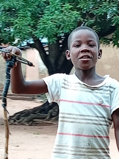 A young, dark-skinned person stands outdoors holding a spear-like object. They are wearing a white t-shirt with horizontal stripes. The person has short hair and is smiling slightly. The spear is long, thin, and wooden, with some metal or dark-colored material wrapped around its lower portion.
Behind the person is a large tree with exposed roots, and a building is partially visible in the background. The ground appears to be dirt or sand.
While the city is mentioned in the prompt, the visual information available in the image is not sufficient to confirm the location.