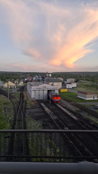 This image shows a view from above of a train yard with multiple railway tracks. A red and silver train is visible on one of the tracks, with its headlights on. The train appears to be stationary. There are several buildings and structures along the train yard, including what look like storage containers and industrial buildings. The surrounding area is covered in green vegetation, suggesting a rural or semi-rural location.
The sky is filled with soft, pastel-colored clouds, indicating either sunrise or sunset. The lighting suggests it's either early morning or late evening. The image is taken from a high vantage point, possibly from a building or a bridge overlooking the train yard.
The word "1976" is clearly visible on the side of one of the larger white buildings. There are also some smaller, more indistinct markings on some of the other structures. The overall mood of the image is one of quiet industry and the beauty of a natural sky at twilight. The image seems to capture a moment of stillness within an operational train yard.