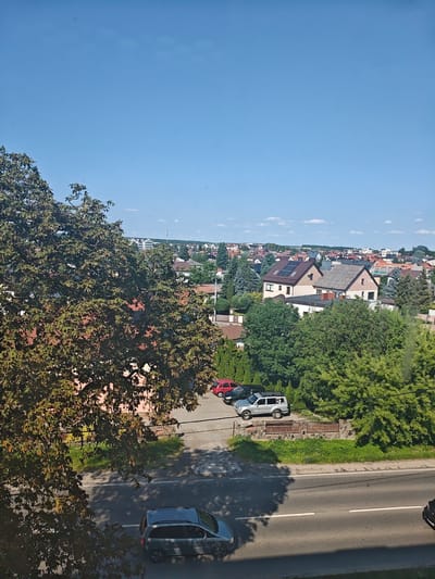 This media file shows an elevated view of a residential area on a clear, sunny day. The sky is a vibrant blue with a few scattered white clouds. In the foreground, a large tree with dense green foliage partially obscures the view. Beyond the tree, a street runs horizontally across the lower part of the image. Several vehicles are visible on the street and in a parking area. A gray minivan is driving on the street, and a silver SUV, a black car, and a red car are parked in a lot.
The scene transitions to a collection of houses with pitched roofs, many of which appear to be residential homes. Some houses have solar panels installed on their roofs. Numerous trees and greenery are interspersed throughout the neighborhood, suggesting a well-established suburban or town setting. In the distance, more buildings and some wind turbines can be seen, indicating the presence of a larger town or city beyond the immediate residential area.
The lighting and clear sky suggest it is daytime, likely late morning or afternoon. The overall atmosphere is peaceful and ordinary, depicting a typical day in a Polish town. There is no visible text within the image. The context provided places the location in Mława, Poland.