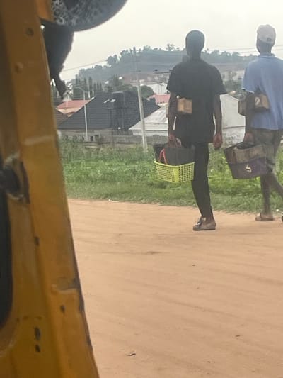 In Galadimawa Village, Nigeria, two individuals walk along a dusty road. The person in the foreground, wearing a black t-shirt and dark trousers, carries a lime green plastic basket filled with dark bags. They are also wearing brown shoes. To their right, a second person, dressed in a blue t-shirt and khaki trousers, carries a dark bag over their shoulder. They are wearing sandals. In the background, residential buildings with dark roofs are visible, interspersed with green vegetation and a hazy sky. The foreground is dominated by the yellow structure of what appears to be a vehicle.