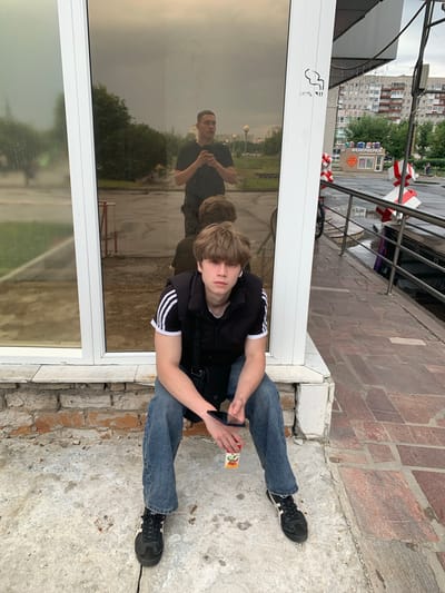 A young person with blond hair is sitting on a concrete ledge in front of a building with large reflective windows. They are wearing a black t-shirt with white stripes on the sleeves, blue jeans, and black athletic shoes. The person is holding a small carton of juice with a picture of fruit on it and a smartphone in their hands.
In the reflection of the window, another person is visible, wearing a black t-shirt and looking towards the camera. The background of the reflection shows trees and buildings, suggesting an outdoor urban environment.
The scene appears to be set in the daytime under an overcast sky. The ground is made of concrete and brick.
The visible text on the building's window reads "GIORGIO". There is also a "no smoking" symbol on the window. In the background, there are buildings and some red and white decorative elements, possibly related to a festival or event. The location is identified as Severodvinsk, Russia.