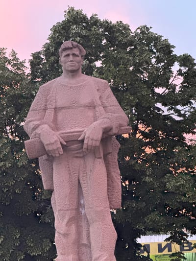 An outdoor, eye-level, full-shot photograph features a large, pink stone statue of a man in the foreground, with a backdrop of lush green trees and a soft pink and blue sky. The statue depicts a stoic figure with a determined expression, wearing a textured tunic and trousers, and holding a rifle horizontally across his chest with both hands. The rifle is positioned in front of his abdomen, with his left arm supporting its weight. The texture of the statue is rough and granular, suggesting it is made of stone or concrete.
Behind the statue, a dense canopy of green trees fills the middle ground, their leaves obscuring much of the background. Through the gaps in the foliage, glimpses of a vibrant pink and pale blue sky can be seen, indicating either sunrise or sunset. The lighting suggests the photograph was taken during the golden hour.
In the lower right corner of the image, a yellow sign with dark blue Cyrillic lettering is partially visible behind the trees and statue. Some of the letters appear to be "М И В". This sign, along with the statue itself, suggests a public space within a city. While the provided information states the location is Orizovo, Bulgaria, this is not visually discernible from the image alone. The overall atmosphere is one of solemnity and possibly historical significance, given the nature of the statue and its public display.