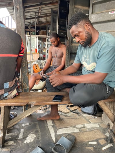 Two men are sitting on a wooden bench. One man wears a light blue shirt with a Lacoste logo and dark pants. The other man is shirtless and wears dark shorts. The man in the shorts is cutting his toenails. A black smartphone and a black bag are on the bench next to the men. A tile floor is visible.