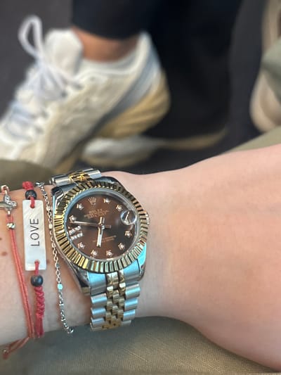 A person's wrist is shown wearing a two-tone stainless steel and gold Rolex Datejust watch with a brown dial and diamond hour markers. The watch is worn above several bracelets: a silver chain, a red braided string bracelet with a white tag that says "LOVE," and a red string bracelet with a silver cross charm. Part of a white and gold sneaker is visible in the background. The person is wearing a green jacket. There is no information in the image that references the city of Alessandria, Italy.