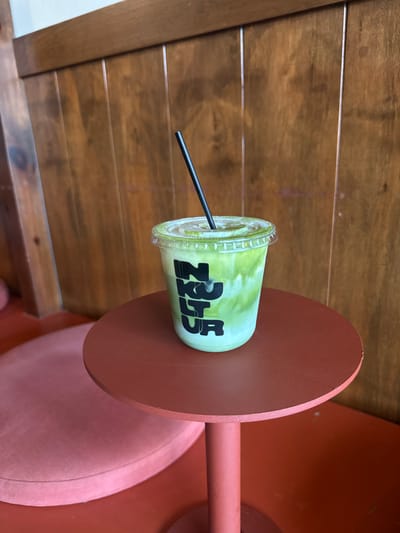 A close-up shot presents a cup of green matcha latte with a black straw, resting on a small, round, terracotta-colored table attached to a coral-colored chair with a plush, pink cushion. The cup is labeled with "ZUZU," the name of the coffee shop. The background features a wooden wall with horizontal panels. This coffee shop is located in San Pedro Garza García, Mexico.