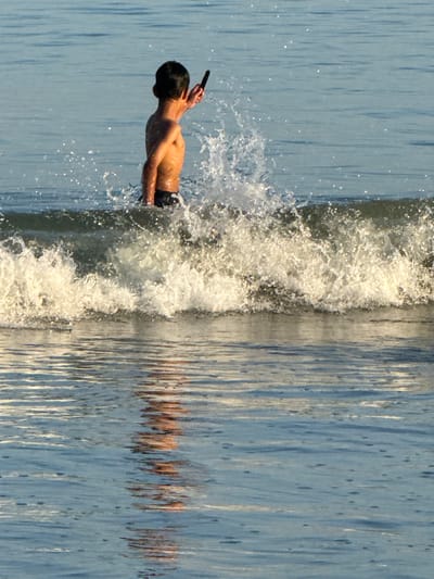 The image, taken in Jimbaran, Indonesia, depicts a shirtless individual, likely a young male, holding a black object to his head. He is positioned horizontally in the frame, seemingly suspended mid-air as if he is diving. Splashes of water surround him. The background consists of what appears to be a vertically-oriented water surface, possibly the ocean, with distorted reflections visible.