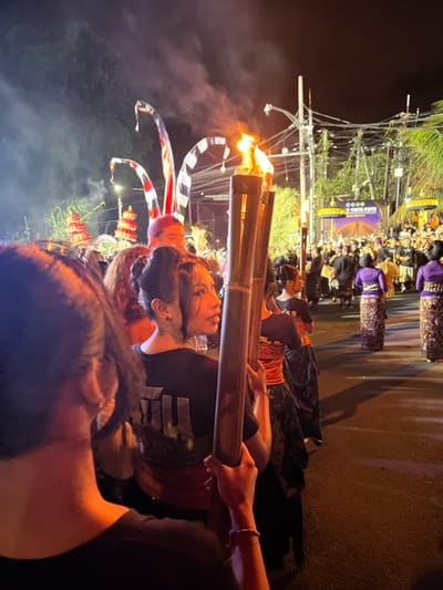 This image captures a nighttime ceremonial procession on a street in Munggu, Indonesia. In the foreground, the blurred left shoulder and back of an individual holding a bamboo torch are visible. Immediately to their right, a young woman with dark hair styled in two buns, wearing a black t-shirt with a light logo and a red sash over a dark patterned sarong, faces right, holding three flaming bamboo torches that emit smoke. Behind her, another participant in an orange top and patterned sarong also carries a torch. Further back, a line of individuals dressed in purple tops and dark patterned sarongs walks away from the viewer. To the left, several tall, curved ceremonial poles with red, white, and black striped fabric, and ornate tiered structures are visible. In the background, a large crowd gathers under artificial lighting, with some people in traditional attire. A prominent banner displays "OOOO GOGO OPEN" over an illuminated graphic of a bridge. Overhead utility lines and streetlights are visible against the dark sky.