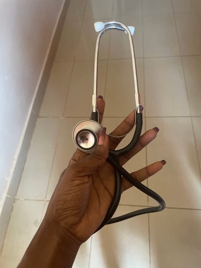 A person's hand, with dark skin and dark nail polish, holds a silver and black stethoscope. The stethoscope has two white ear tips and a metal chestpiece with a black diaphragm. The hand is positioned in the center of the frame, with the stethoscope extending upwards and to the right. The background consists of a tiled floor and a wall. The scene is likely indoors. There is no indication of the specific location within the city of Kakau, Nigeria.