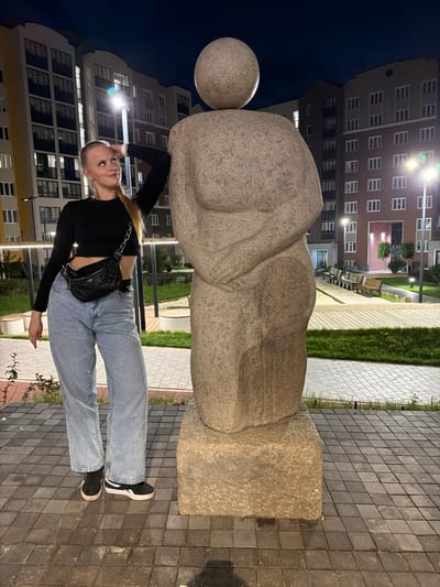 In the city of Pervomaysky, Russia, at night, a young woman with blonde hair pulled into a ponytail stands next to a large stone sculpture. She is wearing a black long-sleeved crop top, light-wash wide-leg jeans, and black sneakers. A black quilted fanny pack is slung across her body. The sculpture, appearing to be abstract or figurative, is made of rough-textured stone and has a spherical element on top. Behind them are modern apartment buildings with numerous windows, some illuminated. The ground is paved with square tiles. Benches and green grassy areas are visible in the background, along with streetlights. The lighting suggests it is evening or nighttime.