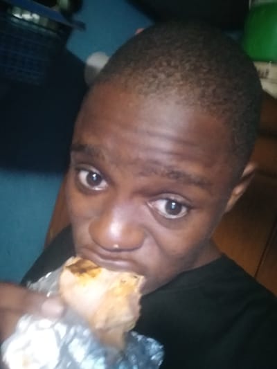 An individual with short, dark hair is holding a food item wrapped in foil near their mouth, presumably about to eat it. The person has brown eyes and dark skin. The background is blurred and shows a blue wall, a dark object that may be a basket, a green object that might be a container and a wooden surface, perhaps a cabinet or door. It is not possible to verify the location as Epe, Nigeria.