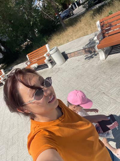 The image shows two people outdoors on a sunny day, likely in Pavlodar, Kazakhstan. In the foreground, a woman is taking a selfie, wearing an orange t-shirt and sunglasses. She has brown hair and is smiling. Her left hand is visible, holding a burgundy wallet. Behind her, a young child wearing a pink baseball cap and a white shirt is also visible, looking up and reaching out. The ground is paved with light-colored tiles, and to the left, a grassy area and trees are present. In the background, there are park benches made of wood and concrete. The lighting suggests it is daytime, with strong shadows indicating bright sunlight. The overall mood appears cheerful and relaxed, possibly during a leisurely outing in a park or public space. No visible text is present in the image.