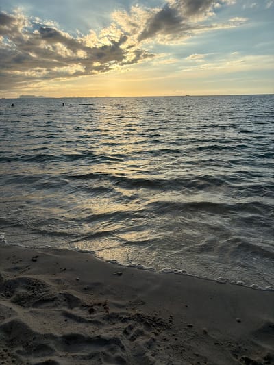 The image depicts a serene sunset scene at the beach in Baan Tai, Thailand. The sky is a canvas of muted blues and greys, illuminated by the soft, golden glow of the setting sun peeking through scattered clouds. The sun's rays cast a shimmering path across the tranquil sea, which gently laps at the sandy shore. In the distance, silhouettes of people can be seen wading in the water, enjoying the peaceful atmosphere. The foreground is dominated by the dark, textured sand of the beach, with the gentle curve of the shoreline marking the boundary between land and sea.