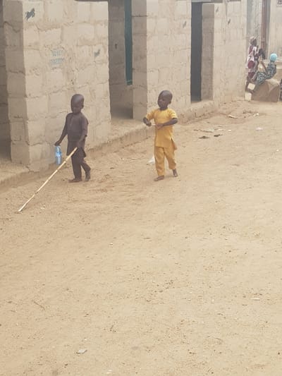 The image depicts an outdoor scene in Dutsinma B, Nigeria. Two young boys are present. One, clad in a yellow outfit, stands on the dirt ground. The other, wearing a darker outfit, stands near the edge of a rough concrete structure holding a stick and a water bottle. In the background, a group of people are clustered near a building. The ground is sandy and sparsely littered.