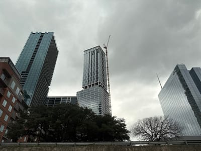 The image captures several modern skyscrapers under a gray, overcast sky. One building is under construction with a crane visible atop it. A tall, glass building reflects the sky. A shorter brick building, labeled "Camden," is also present. Bare branches of a tree frame the foreground, adding a natural element to the urban scene. A low wall or barrier is at the bottom edge of the frame.