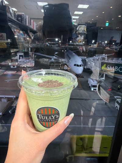 The image displays a Tully's Coffee drink, likely a smoothie, held in a person's hand. The drink is green and has chocolate shavings on top. The location is at Narita International Airport in Narita, Japan. This is inferred from the exterior view reflected in the window behind the drink. The view shows an aircraft and airport infrastructure, including signage that suggests it's a major international hub, namely Narita International Airport. The reflections in the window also indicate an indoor setting, like a terminal or lounge inside the airport.