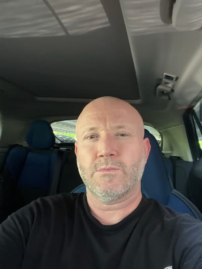 A close-up photograph depicts a Caucasian male with a shaved head, graying beard, and light skin, situated in what appears to be the driver's seat of a vehicle. He is wearing a black shirt. The interior of the vehicle is visible, including the dashboard and part of the passenger seat, which is blue. Outside the car window, a blurred view of greenery suggests a roadside or parking area. There is no information to ascertain that the photo was taken in Sint-Niklaas, Belgium.