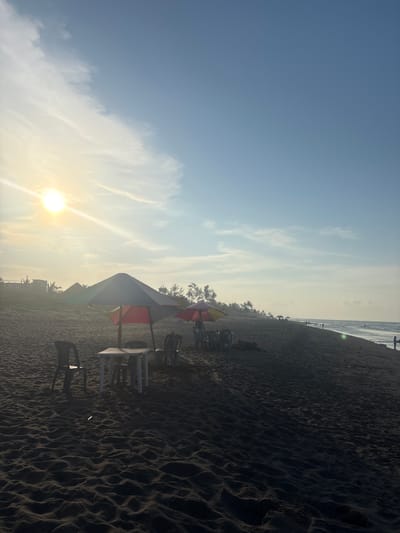 The image depicts a view of the beach in Casitas, Mexico, under a bright, hazy sun. The sky is a clear blue with some wispy white clouds. The sun, a bright disc, is positioned in the upper left quadrant of the image, casting a warm glow and creating a lens flare effect. The foreground is dominated by dark, sandy beach with visible ripples and textures.

In the middle ground, several beach chairs and tables are arranged, some under colorful umbrellas, indicating a seating area for beachgoers. One umbrella is predominantly gray and red, while another is a smaller, multi-colored umbrella with yellow and red sections. The tables appear to be small, white plastic tables. Several plastic chairs are placed around these tables.

Further down the beach, a line of palm trees and other foliage is visible against the horizon, with some structures that appear to be buildings peeking through the trees on the far left. The ocean stretches into the distance on the right side of the image, with small waves lapping at the shore. A few people are sparsely scattered along the beach and in the water, suggesting a relatively uncrowded scene. The overall impression is one of a calm, late afternoon or early morning beach environment.