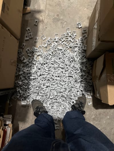 The scene appears to be inside a warehouse or storage area, with boxes stacked around a large pile of small, gray plastic clips scattered on the concrete floor. A person wearing jeans and gray sneakers is standing near the pile of clips. The boxes appear to be holding the clips. The location of the scene is Campana, Argentina.