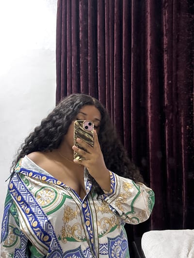 A woman with long, dark, curly hair is taking a selfie with her iPhone, partially obscuring her face. She is wearing a white, blue, and green patterned blouse. The background consists of a dark burgundy textured wall. The location in the city of Port Harcourt, Nigeria cannot be determined from the image.