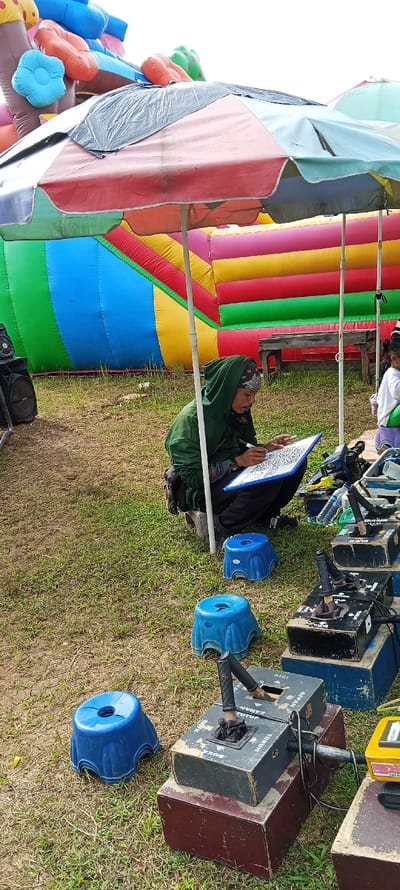 An outdoor scene in Bandasari, Indonesia, depicts a person squatting under a large, multi-paneled umbrella providing shade. The individual wears a dark green hooded jacket, dark pants, and a bandana, and is actively drawing or writing on a white clipboard with a pen. To their immediate left is a black speaker. To their right, a collection of equipment includes two small blue plastic stools on the grass, a stack of dark wooden or metal control boxes with levers and wires, and a yellow electronic device. One control box displays Indonesian text such as "KIRI," "DEPAN," "KANAN," "TUTUP," and "BUKA." A clear plastic water bottle rests near the equipment. In the background, a large, multi-colored inflatable structure (red, yellow, green, blue, pink, with orange and light blue elements) dominates, suggesting a recreational event. A rustic wooden bench or table is visible behind the equipment, and a small child is partially visible on the far right. The ground consists of sparse green grass and visible dirt. The sky above is overcast and white.