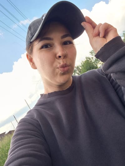 A young woman wearing a dark grey baseball cap and a dark grey sweatshirt is taking a selfie outdoors. She is making a kissing face with her lips puckered. She is holding the brim of her cap with her right hand. The background consists of a bright blue sky with fluffy white clouds, some trees, and green grass. In the distance, there appears to be a building and some electrical wires. The lighting suggests it is daytime. The image is taken from a low angle, looking up at the woman.