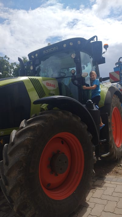 A young boy in a blue t-shirt and dark shorts is climbing into a large, green and black tractor. The tractor has the brand "CLAAS" and the model "830 AXION" visible on its side. The boy is holding onto a handle and has one foot on a step. The tractor has large, rugged tires with orange rims. The background shows a cloudy blue sky and some trees. The scene is set on a paved surface, possibly a farm or a rural area. The overall impression is one of a child interacting with agricultural machinery. The image is taken from a low angle, emphasizing the size of the tractor.