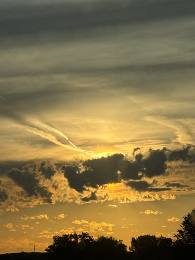 The image captures a dramatic sunset over the city of Enevo, Bulgaria. The sky is filled with a gradient of colors, transitioning from a deep gray at the top to warm oranges and yellows near the horizon. Several layers of clouds are present, with dark, ominous formations contrasting against the brightly lit lower clouds. A prominent contrail streaks across the sky, illuminated by the setting sun. The foreground is silhouetted against the vibrant sky, showing the dark outlines of trees and what appear to be power lines or transmission towers on the horizon. The overall impression is one of a powerful and atmospheric natural display.