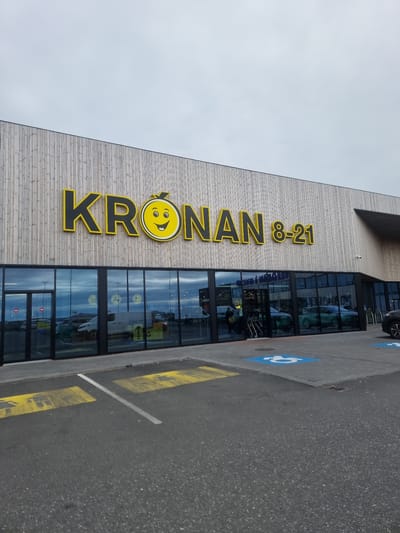 The image shows the exterior of a Krónan supermarket in Njarðvík, Iceland, on an overcast day. The building has a modern facade with a light-colored wooden exterior and large glass windows. The prominent yellow and black Krónan logo, featuring a smiley face within a lemon, is displayed above the entrance. The sign also indicates the store's operating hours as "8-21". Through the glass doors, several shopping carts and people are visible inside the store. The parking lot in front of the store has designated parking spots, including disabled parking spaces marked with blue wheelchair symbols. There are vehicles parked in the lot, including vans and cars. The ground in the parking area is paved asphalt with yellow painted lines and markings.