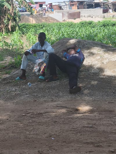 Two young males are in the foreground of the image, situated on a pile of gravel in Kwamba, Nigeria. The male on the left is seated upright, wearing a light blue traditional tunic and trousers. He is holding a clear plastic bag containing toy cars. The male on the right is lying down on the gravel, resting his head on his folded arms. He is wearing a dark blue t-shirt with lettering and black trousers. Behind them, a field of green crops, possibly corn, stretches into the distance, with a backdrop of buildings and structures, some appearing to be under construction with stacks of concrete blocks. The ground is covered in gravel and dirt, with some scattered leaves and debris. Sunlight casts shadows, indicating an outdoor setting.